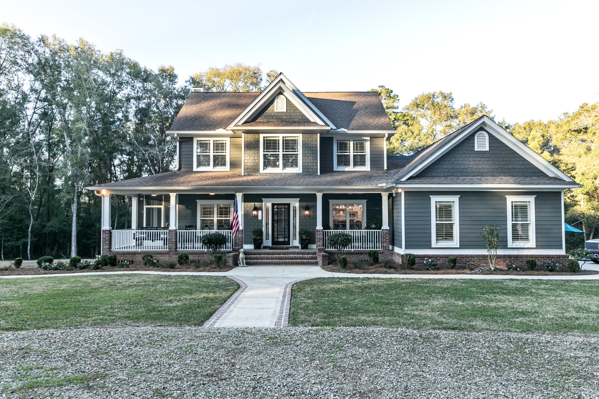 Front view of a large two story blue gray house with wood and vinyl siding with an expansive porch, sidewalk and manicured lawn.